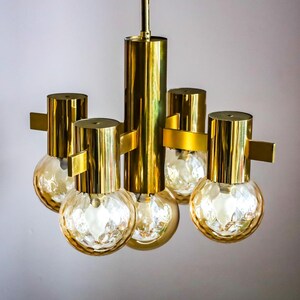 May include: A gold-toned chandelier with five light bulbs. The bulbs are clear glass with a textured surface and emit a warm glow. The chandelier has a modern design with a central column and four arms extending out.