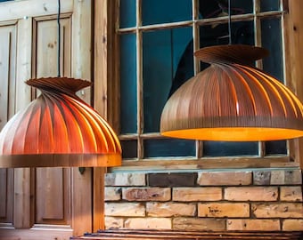May include: Two wooden pendant lights with a unique, layered design. The lampshades are a warm brown color, with a spiral top and a rounded, ribbed bottom. The lights hang in front of a window and brick wall.