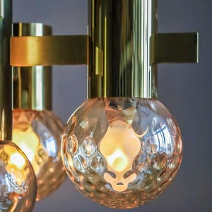 May include: A brass chandelier with six light bulbs. The bulbs are encased in clear glass with a textured surface. The chandelier has a modern design.