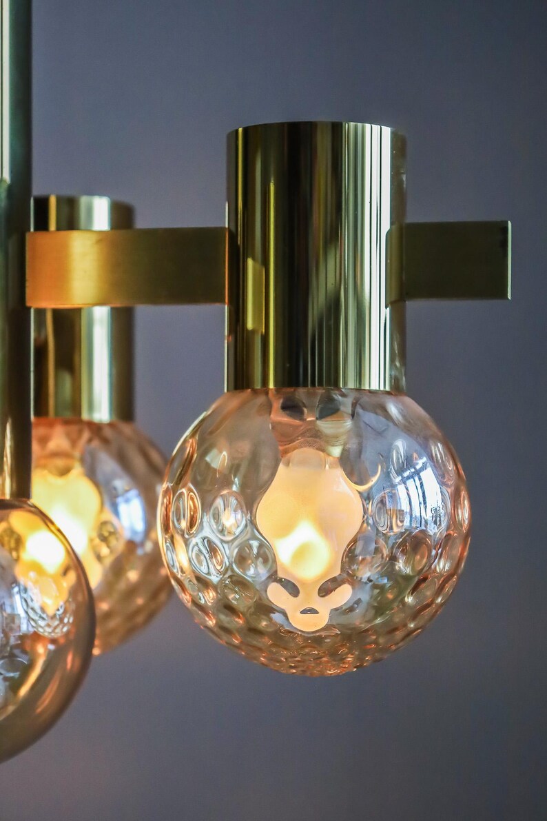 May include: A brass chandelier with six light bulbs. The bulbs are encased in clear glass with a textured surface. The chandelier has a modern design.
