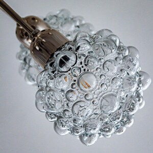 May include: Close-up of a clear glass pendant light fixture with a bubbled texture. The light fixture is attached to a silver metal rod.
