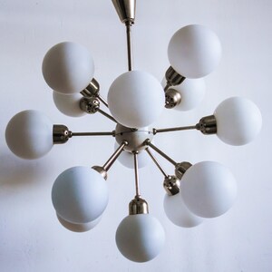 May include: A modern, mid-century style chandelier with a brass finish and ten white frosted glass globes. The chandelier has a starburst design with the globes extending outward from a central point.