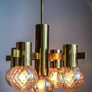 May include: A brass chandelier with five frosted glass globes. The chandelier has a simple design with a central rod and four arms extending out to hold the globes. The globes are round and have a textured surface.