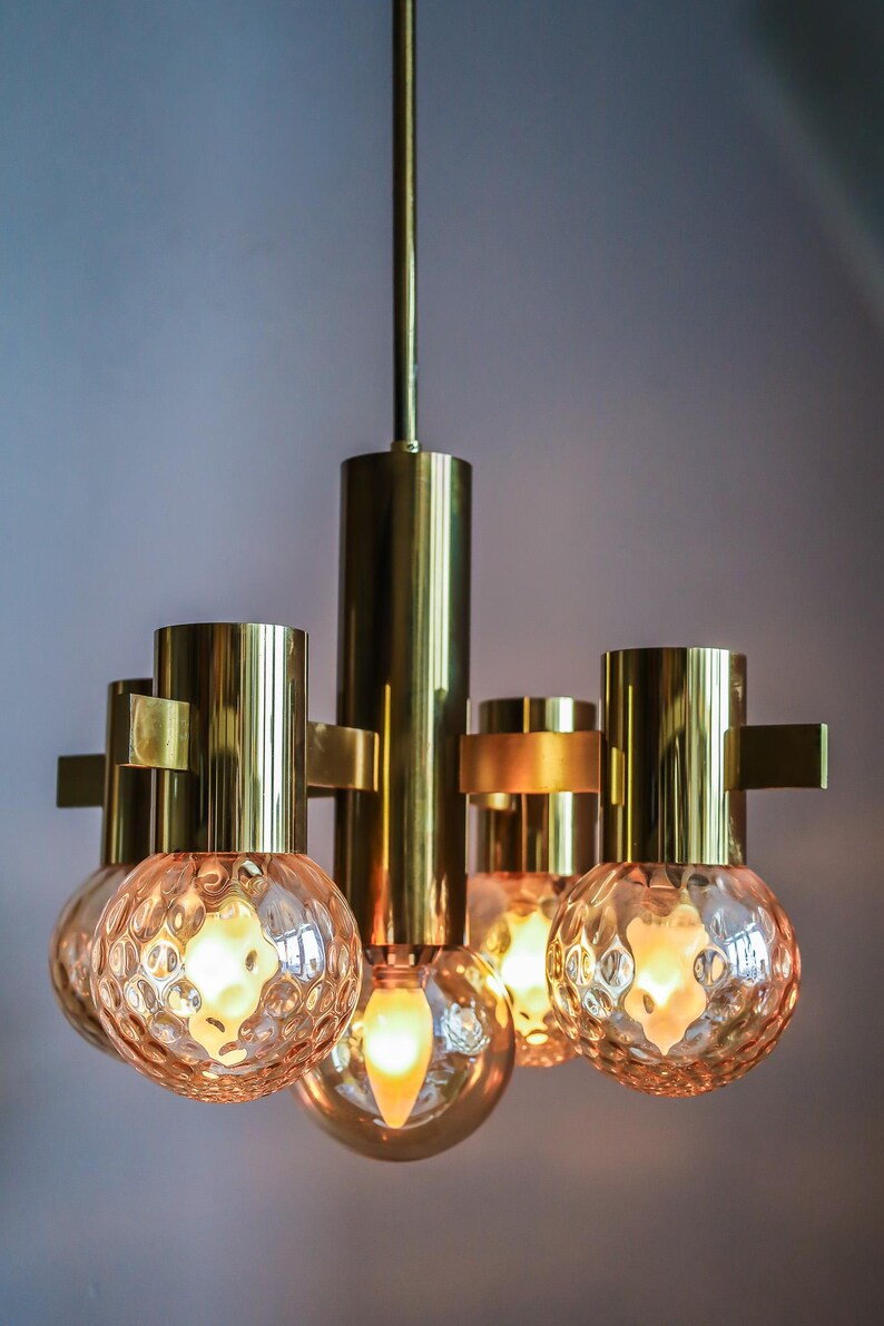 May include: A brass chandelier with five frosted glass globes. The chandelier has a simple design with a central rod and four arms extending out to hold the globes. The globes are round and have a textured surface.