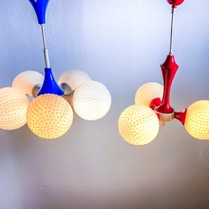 Może przedstawiać: Dwa nowoczesne żyrandole sufitowe z białymi kloszami w kształcie kuli. Żyrandol po lewej stronie ma niebieską podstawę i pięć kloszy, podczas gdy żyrandol po prawej stronie ma czerwoną podstawę i cztery klosze.