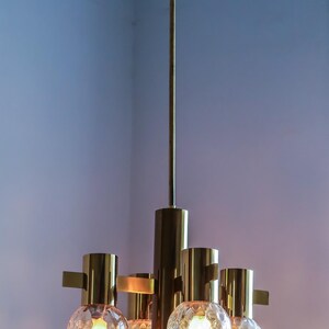 May include: A brass and glass chandelier with five light bulbs. The chandelier has a long, thin stem and five arms that extend out from the stem. Each arm has a round, textured glass globe that surrounds a light bulb. The chandelier is hanging from the ceiling.