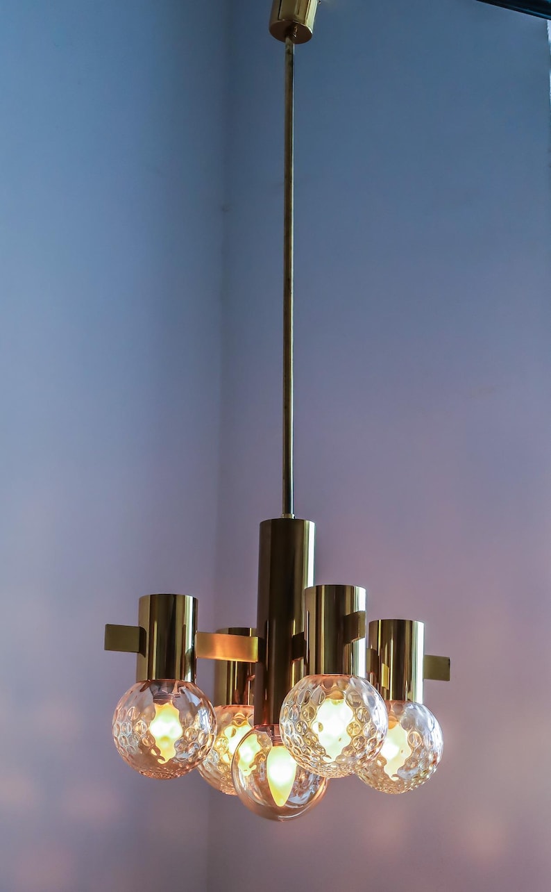 May include: A brass and glass chandelier with five light bulbs. The chandelier has a long, thin stem and five arms that extend out from the stem. Each arm has a round, textured glass globe that surrounds a light bulb. The chandelier is hanging from the ceiling.