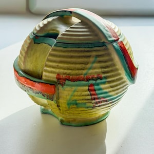 May include: A ceramic lidded container with a handle, featuring a ribbed texture and a vibrant color palette. The base is yellow, accented with red, green, and blue brushstrokes. The handle and rim are also painted with the same colors.