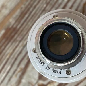 K&ouml;nnte beinhalten: Nahaufnahme eines Canon Kameraobjektivs. Das Objektiv ist silberfarben mit schwarzem Innenraum und einer Glaslinse. Die Worte "MADE BY CANON" sind auf dem silbernen Rand aufgedruckt. Das Objektiv liegt auf einer Holzoberfl&auml;che.