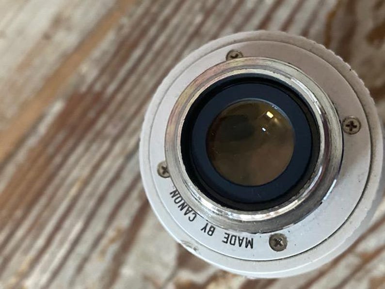 K&ouml;nnte beinhalten: Nahaufnahme eines Canon Kameraobjektivs. Das Objektiv ist silberfarben mit schwarzem Innenraum und einer Glaslinse. Die Worte "MADE BY CANON" sind auf dem silbernen Rand aufgedruckt. Das Objektiv liegt auf einer Holzoberfl&auml;che.