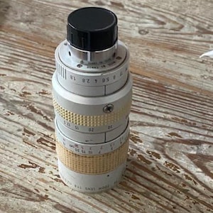 K&ouml;nnte beinhalten: Ein Canon Zoomobjektiv, Modell 118, mit schwarzer Kappe und silber-goldfarbenem Geh&auml;use. Das Objektiv hat verschiedene Messmarkierungen und einen strukturierten Griff. Das Objektiv ist f&uuml;r die Fotografie konzipiert.