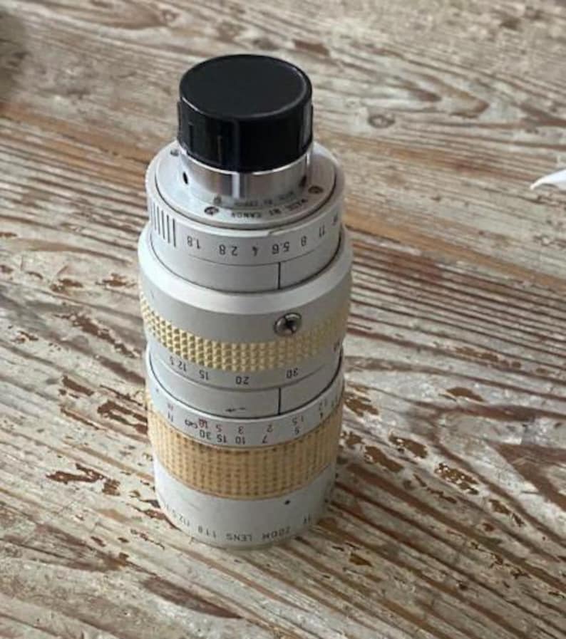 K&ouml;nnte beinhalten: Ein Canon Zoomobjektiv, Modell 118, mit schwarzer Kappe und silber-goldfarbenem Geh&auml;use. Das Objektiv hat verschiedene Messmarkierungen und einen strukturierten Griff. Das Objektiv ist f&uuml;r die Fotografie konzipiert.
