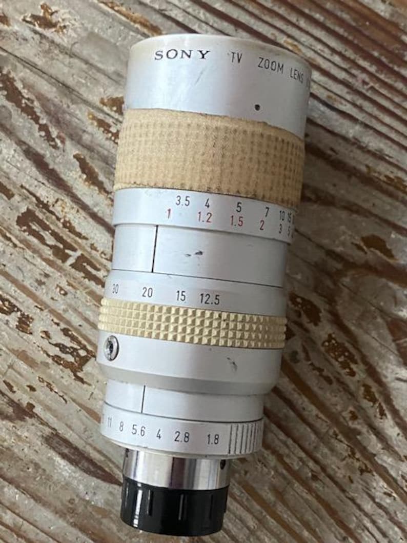 K&ouml;nnte beinhalten: Ein Vintage Sony TV Zoom-Objektiv mit silbernem Geh&auml;use und beigefarbenen und wei&szlig;en strukturierten Griffen. Das Objektiv hat verschiedene numerische Markierungen und eine schwarze Objektivkappe. Der Text "SONY TV ZOOM LENS" ist sichtbar.