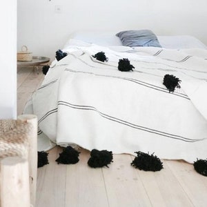 May include: White bed with a black and white striped throw blanket with black pom poms. The bed is in a white room with a small wooden side table and a woven light fixture.