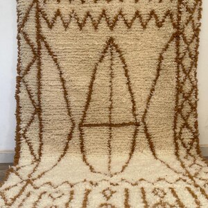 May include: Cream-coloured rug with a brown geometric pattern. The rug features a repeating design of diamonds, triangles, and other shapes. The rug is made of a soft, textured material.