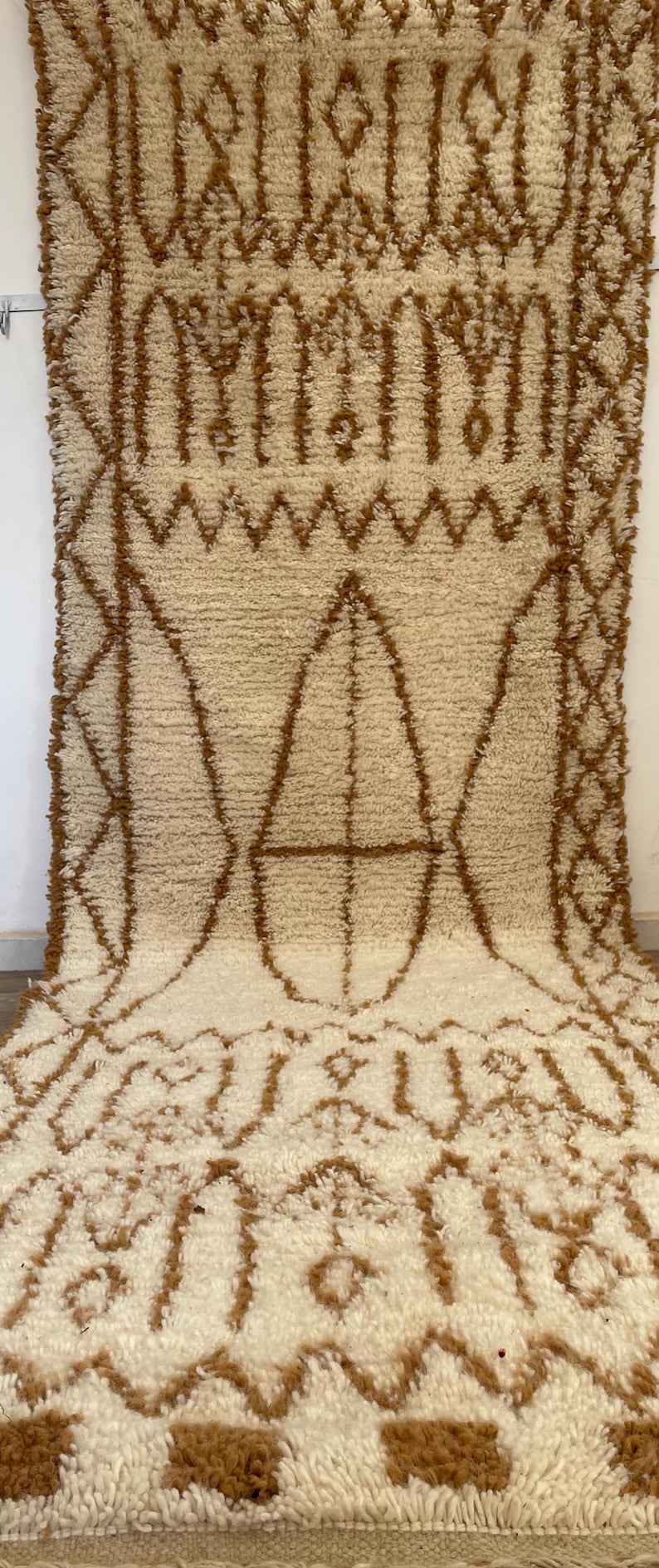 May include: Cream-coloured rug with a brown geometric pattern. The rug features a repeating design of diamonds, triangles, and other shapes. The rug is made of a soft, textured material.