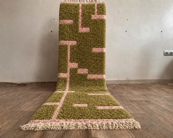 May include: An olive green rug with a pink geometric design. The rug has a shaggy texture and a cream fringe. It is displayed against a neutral wall and wooden floor. The rug's dimensions are not visible.
