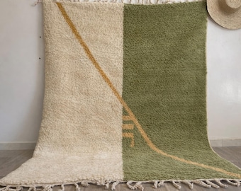May include: A large, rectangular rug with a two-tone design. One half is a creamy white, the other is olive green. A diagonal gold line runs across the rug. The rug has a thick, textured pile and fringe along the bottom edge. A straw hat hangs on the wall.