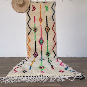 以下が含まれることがあります： 緑、オレンジ、ピンク、青、黒の色合いのカラフルな幾何学模様が施された白いモロッコの絨毯。フリンジの縁取りがあり、ウール製です。