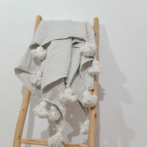 May include: A white and grey striped throw blanket with white pom-poms hanging from the edges. The blanket is draped over a wooden ladder.