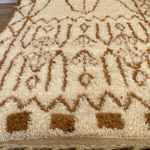 May include: A cream-coloured rug with a brown geometric pattern. The rug has a textured, plush surface and a fringe along the bottom edge. The design features repeating shapes and lines.