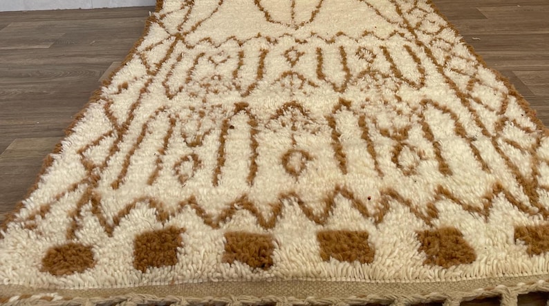 May include: A cream-coloured rug with a brown geometric pattern. The rug has a textured, plush surface and a fringe along the bottom edge. The design features repeating shapes and lines.