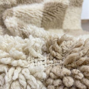 May include: Close-up of a textured, cream-colored rug with a checkered pattern in beige. The rug has a thick, shaggy pile and visible woven backing. The fibers are knotted and create a soft, inviting texture.