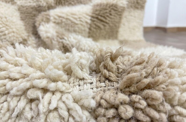 May include: Close-up of a textured, cream-colored rug with a checkered pattern in beige. The rug has a thick, shaggy pile and visible woven backing. The fibers are knotted and create a soft, inviting texture.