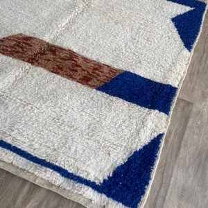 May include: A white rug with blue and brown geometric patterns. The rug has a fluffy texture and a fringe border.