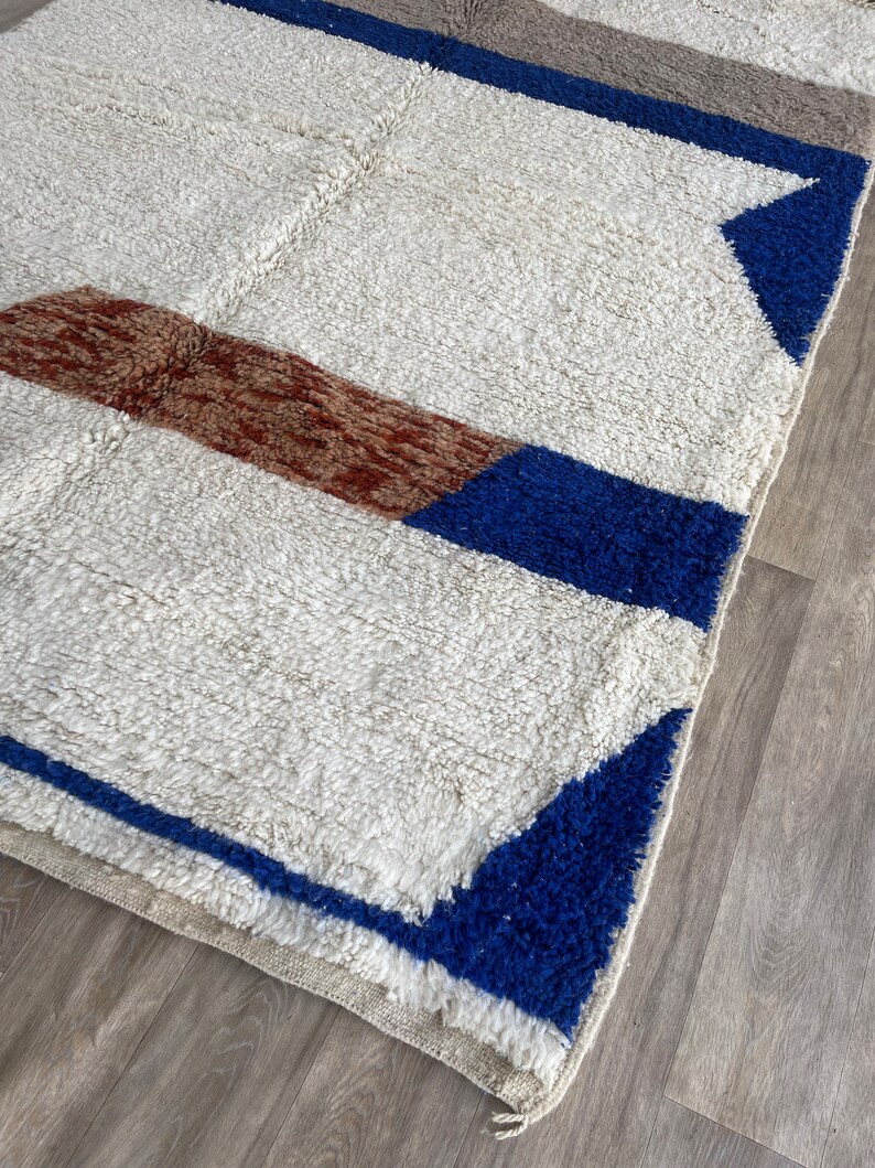 May include: A white rug with blue and brown geometric patterns. The rug has a fluffy texture and a fringe border.