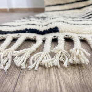May include: A thick, cream-colored rug featuring horizontal black stripes and a textured, shaggy pile. The edge of the rug is finished with long, braided cream-colored fringe tassels that extend outward from the main body of the textile.