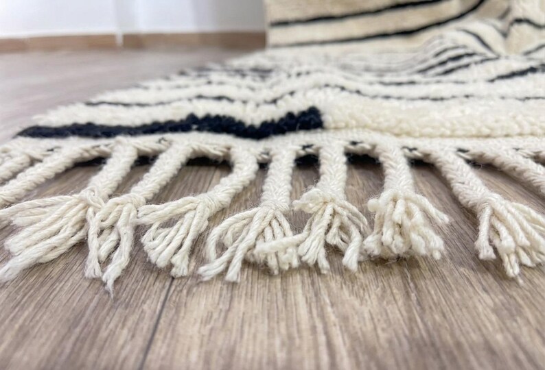 May include: A thick, cream-colored rug featuring horizontal black stripes and a textured, shaggy pile. The edge of the rug is finished with long, braided cream-colored fringe tassels that extend outward from the main body of the textile.
