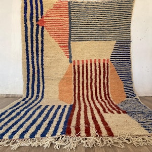 May include: A hand-woven rug with a geometric pattern in shades of blue, red, orange, and beige. The rug has a fringe border.