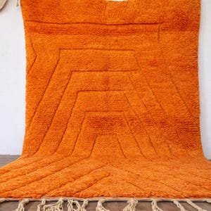 May include: Bright orange, geometric patterned rug with a textured, plush surface. The rug features a central design of layered, angular shapes and a fringe along the bottom edge. The rug is displayed against a white wall.