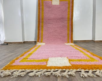 Puede incluir: Una alfombra de pasillo rosa y naranja con un patrón geométrico. La alfombra tiene flecos blancos.