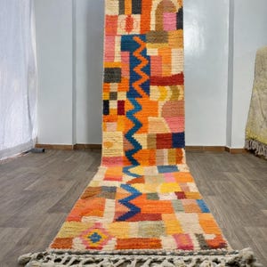 Könnte beinhalten: Ein langer, rechteckiger, farbenfroher Teppich mit geometrischem Muster. Der Teppich weist eine Vielzahl von Farben auf, darunter Orange, Blau, Rosa und Gelb. Der Teppich hat ein Zickzack-Design in der Mitte und Quasten an den Enden. Der Teppich ist etwa 3 Meter lang.