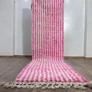 May include: A pink and white striped rug with a textured, shaggy appearance. The rug has a rectangular shape with a fringe along one edge. The vertical stripes alternate between pink and white. The rug is displayed on a wooden floor.