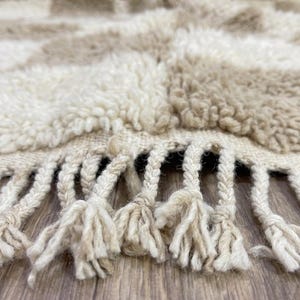 May include: Close-up of a beige and cream-colored rug with a checkered pattern. The rug has a thick, textured pile and a fringe of knotted tassels along the bottom edge. The rug appears to be made of natural fibers.
