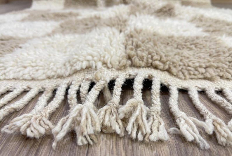 May include: Close-up of a beige and cream-colored rug with a checkered pattern. The rug has a thick, textured pile and a fringe of knotted tassels along the bottom edge. The rug appears to be made of natural fibers.