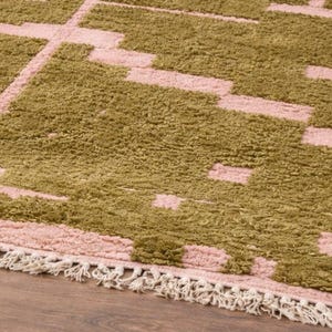 Puede incluir: Una alfombra rectangular con un patr&oacute;n geom&eacute;trico en verde oliva y rosa claro. La alfombra tiene un borde con flecos de color crema y est&aacute; colocada sobre un suelo de madera.