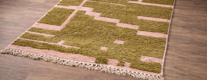 Puede incluir: Una alfombra rectangular con un patr&oacute;n geom&eacute;trico en verde oliva y rosa claro. La alfombra tiene un borde con flecos de color crema y est&aacute; colocada sobre un suelo de madera.