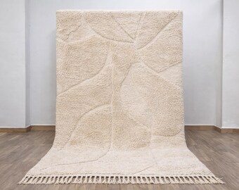 May include: A large, rectangular beige rug with a textured, abstract design. The rug features a series of curved, raised patterns across its surface. The bottom edge has a fringe detail. The rug is displayed against a white wall.