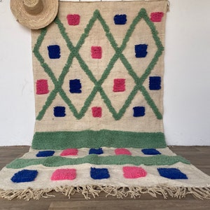 Op de afbeelding: Een wit tapijt met een geometrisch patroon van groene, roze en blauwe vierkanten. Het tapijt heeft een franje rand.