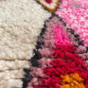 Op de afbeelding: Close-up van een kleurrijk, handgeweven tapijt met een geometrisch patroon. Het tapijt heeft tinten roze, rood, oranje en wit.