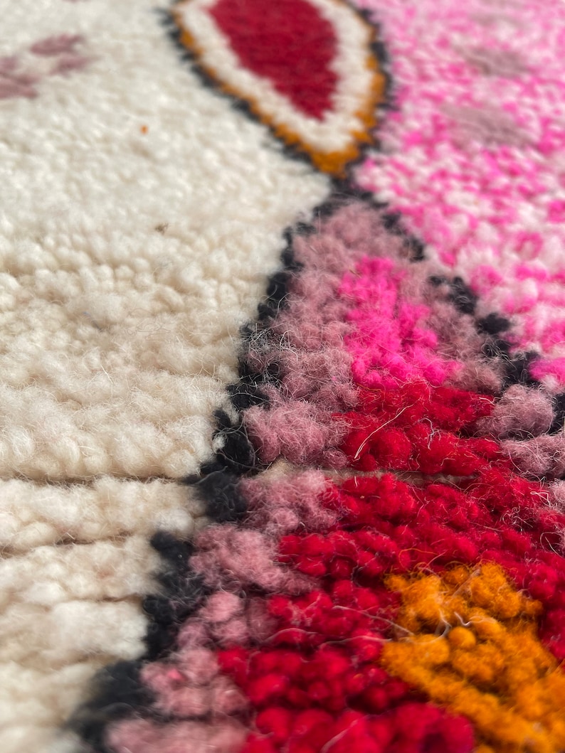 Op de afbeelding: Close-up van een kleurrijk, handgeweven tapijt met een geometrisch patroon. Het tapijt heeft tinten roze, rood, oranje en wit.