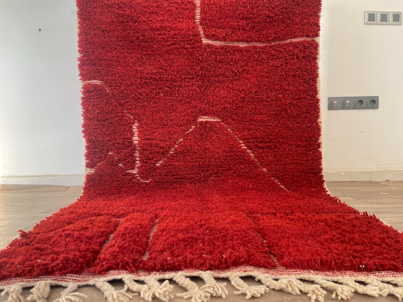 May include: A vibrant red, textured rug with a geometric design. The rug features a mountain-like pattern in white, with a fringe along the bottom edge. The thick pile suggests a soft, comfortable texture, suitable for a living room or bedroom.