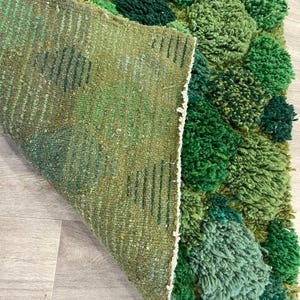 May include: A green rug with a textured, tufted design. The rug features various shades of green, with a raised, three-dimensional pattern. The underside of the rug is visible, showing a woven construction with green and white threads. The rug is on a light-coloured wooden floor.