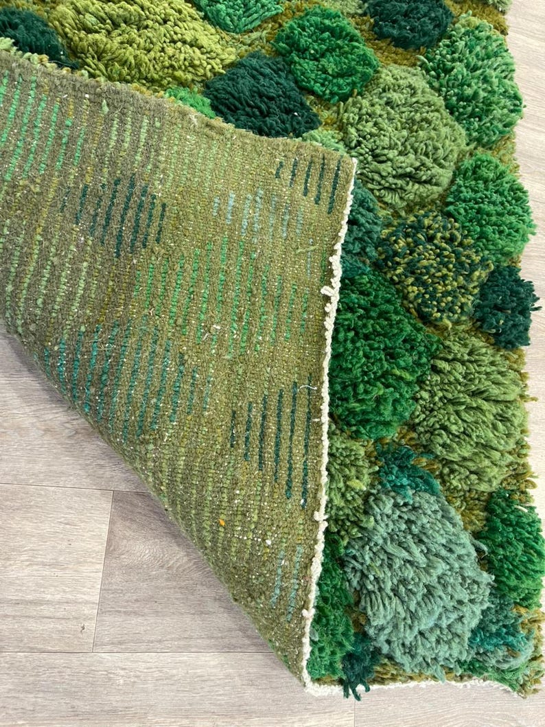 May include: A green rug with a textured, tufted design. The rug features various shades of green, with a raised, three-dimensional pattern. The underside of the rug is visible, showing a woven construction with green and white threads. The rug is on a light-coloured wooden floor.