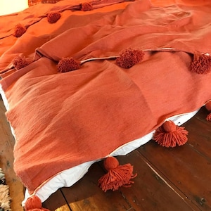 Puede incluir: Una manta de color terracota con ribete blanco y pompones en los bordes.