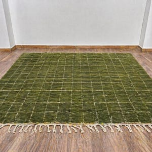 May include: A rectangular green rug with a grid pattern and cream-colored fringe. The rug is made of a textured material and is placed on a wooden floor. The grid pattern is created by white lines.